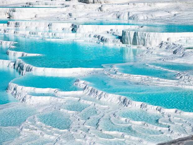 Terraced turquoise pools cascade down natural white stone formations, creating a layered effect. The area appears arid and expansive, highlighting the contrast between the water and rock. There's no text present.